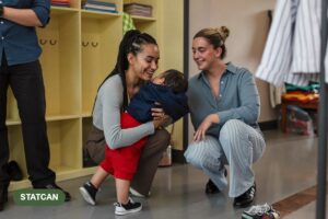 A parent crouches to hug her toddler goodbye at a child care centre cubby area while an educator waits nearby.