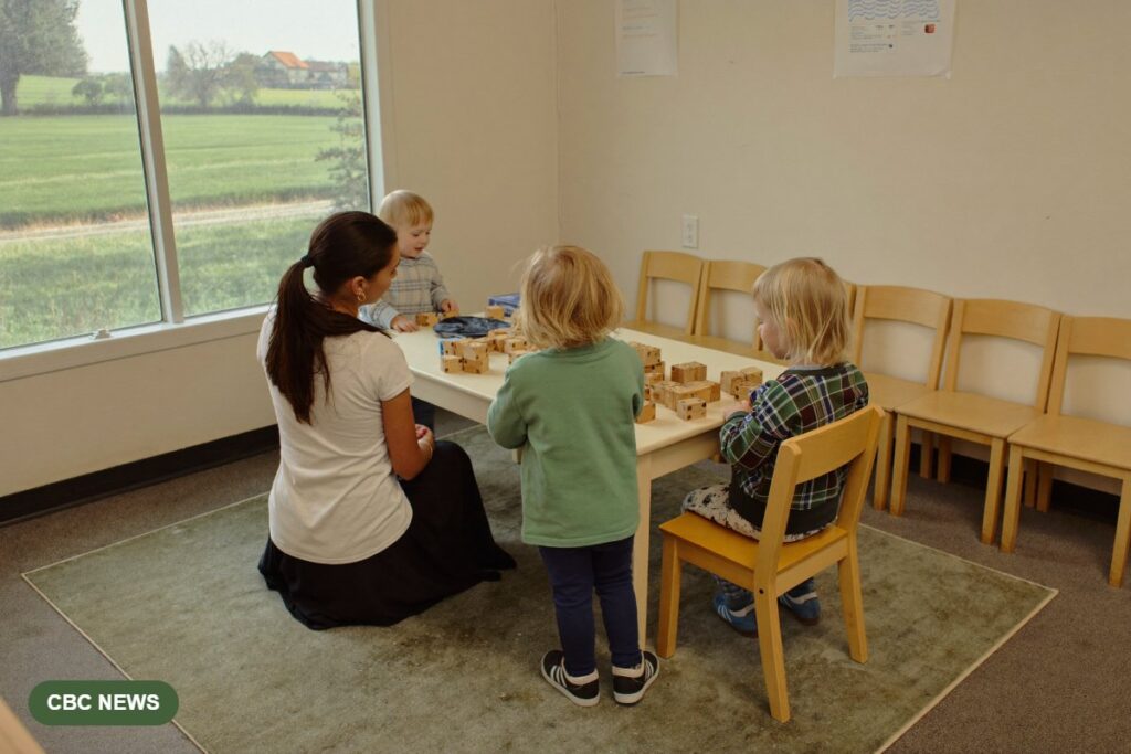 Children play with wooden blocks beside an educator in a small rural child care room, with a row of empty chairs along the wall.