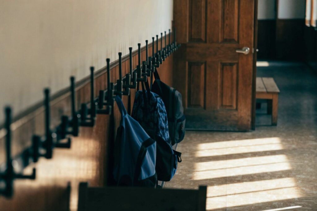 An empty school corridor in late afternoon, with backpacks hanging on a low row of coat hooks at child height.