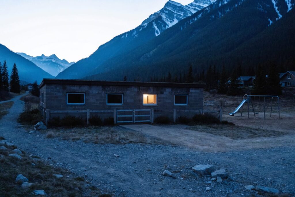 A small single-storey child care building at dusk below snow-covered mountain peaks, with one window lit.